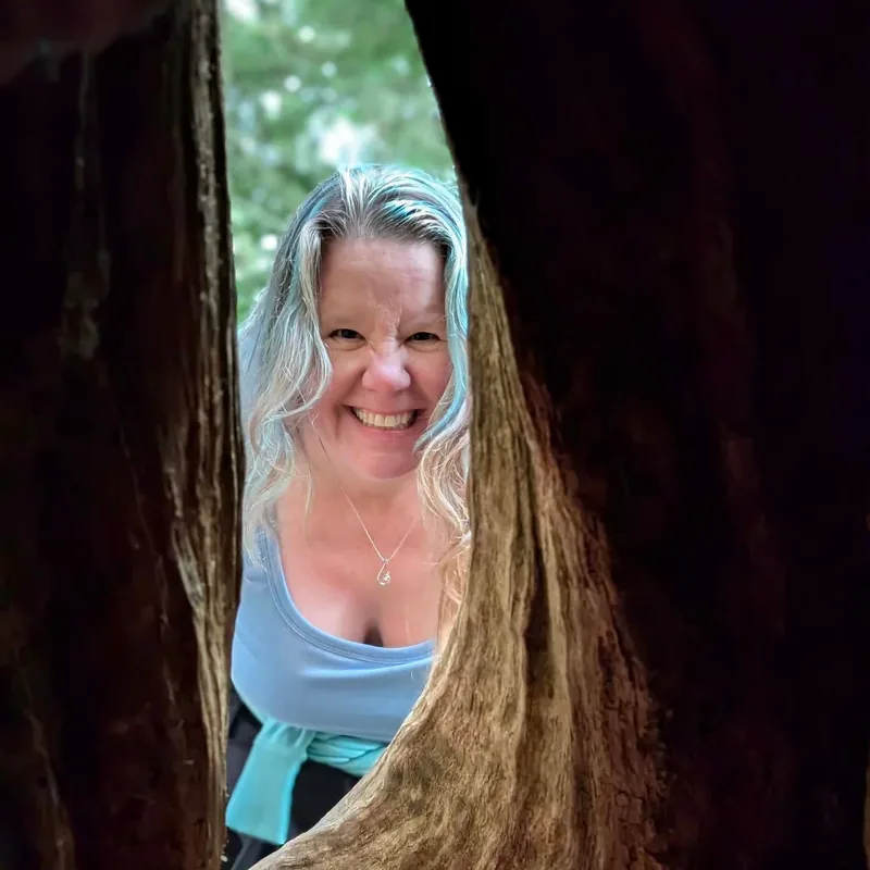 Richelle, grinning through a gap between two redwood trunks